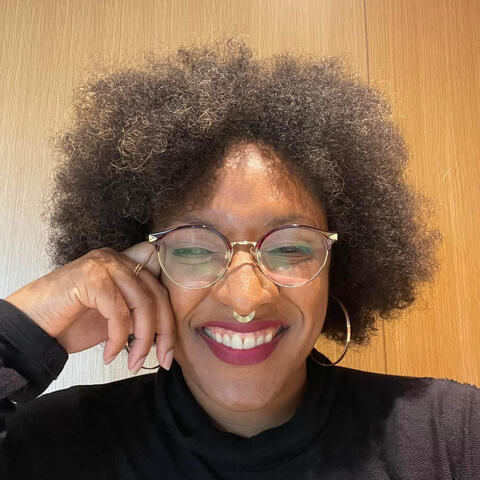 Joya | AI Prompt Wizard | Bomb-Ass Prompts Only Joya Jones, a radiant Black woman with a voluminous afro, wears stylish gold-rimmed glasses, a septum ring, and a black turtleneck. Resting her head on her hand, she smiles confidently, exuding intelligence, creativity, and effortless charm.